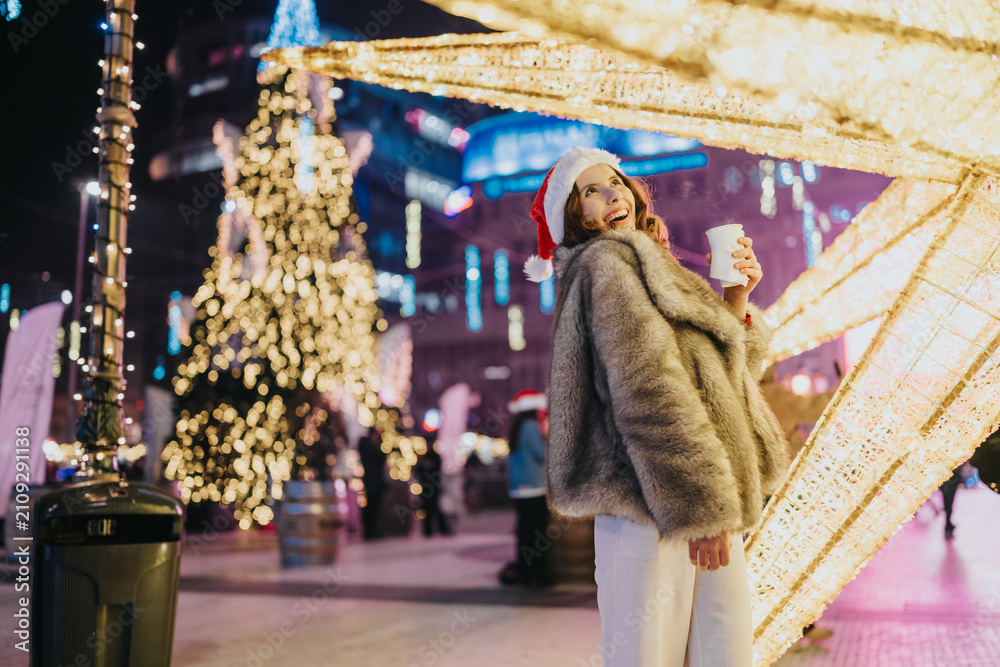 Fototapeta premium Woman wearing a Santa hat and fur coat smiles while holding a warm drink amid holiday lights. Cheerful scene with illuminated decorations and a Christmas tree at a nighttime market.