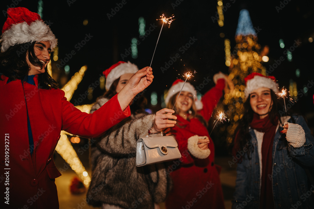 Fototapeta premium Four girls wearing Santa hats hold lit sparklers and smile at night. Friends celebrate outdoors amid festive lights and holiday decorations.