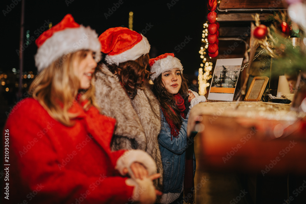 Fototapeta premium Three girls wearing Santa hats browse a festive outdoor Christmas market stall. They look at holiday treats and decorations under warm market lights.