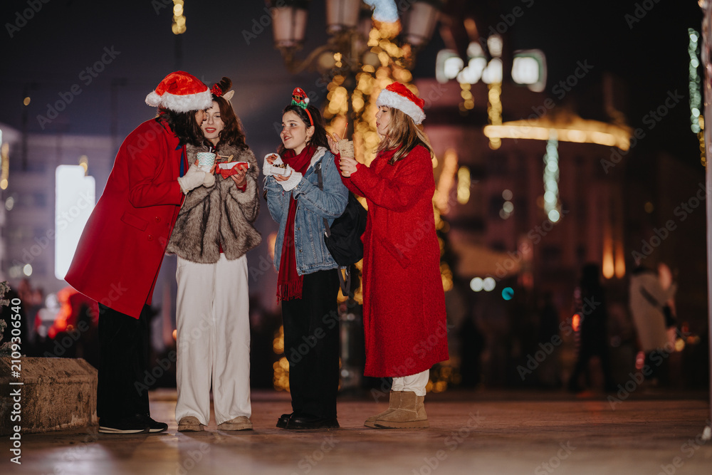 Fototapeta premium Four girls wearing Santa hats share snacks and drinks on a decorated city street at night. Friends laugh and enjoy festive treats together in a winter evening urban setting.