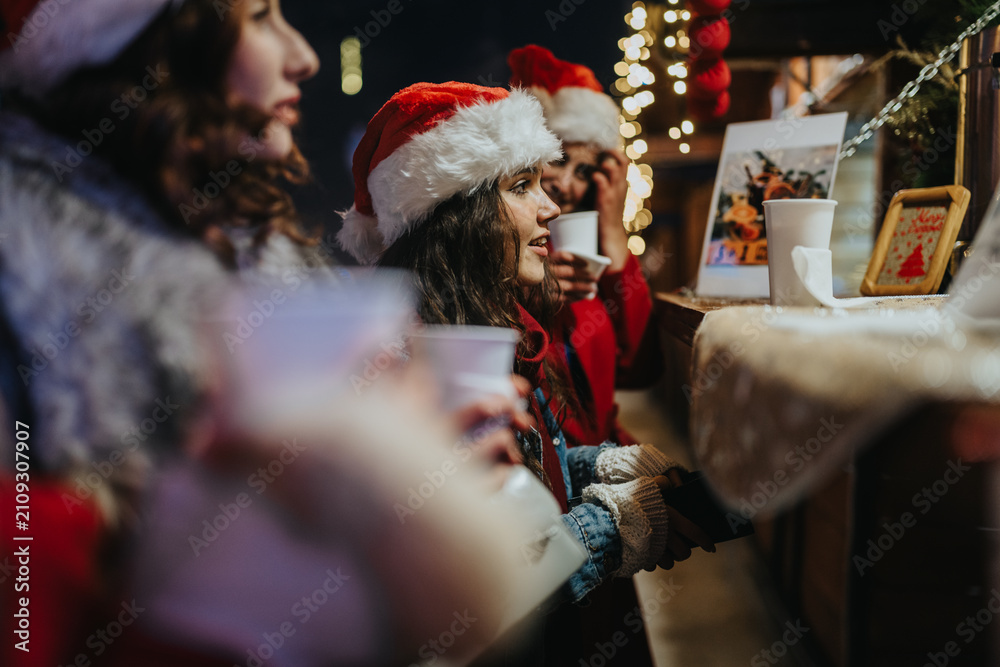 Fototapeta premium Girls wearing Santa hats hold hot cups while smiling at a decorated market counter. A festive outdoor Christmas market scene with warm lights and holiday atmosphere.