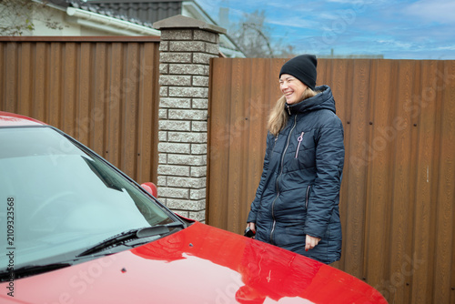 Foto Happy Smiling Mid-Adult Woman in Warm Winter Jacket Standing Near Red Car Outdoo
