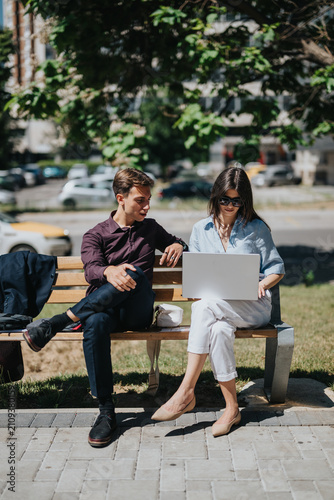 Young colleagues collaborate on a laptop while seated on an outdoor bench. Casual business meeting in a city park with remote work and teamwork.