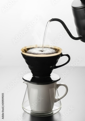 Hot water is being carefully poured over dark ground coffee inside a ceramic dripper resting on a white mug and clear glass server, routine, preparation, home