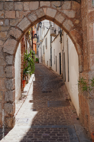 Portalet de la Judería, a medieval stone gateway leading into the historic Jewish Quarter of Sagunto, Valencia, Spain. A picturesque cobbled street preserves the charm and heritage.