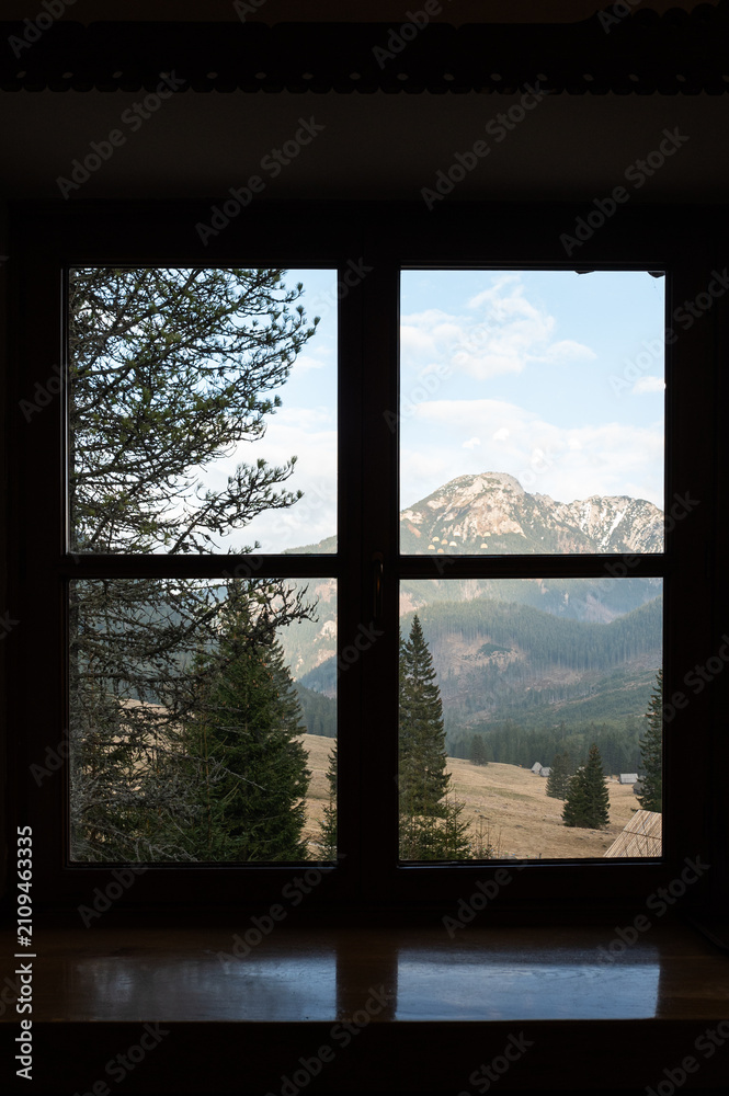 Obraz premium Scenic Mountain View Framed Through a Dark Wooden Cabin Window. A cozy view looking out through a dark wooden window frame towards a beautiful mountain landscape
