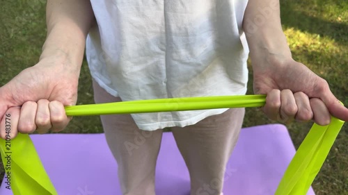 Woman holding green resistance band with both hands on purple yoga mat outdoors on grass. fitness, exercise, strength, training, wellness