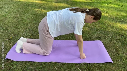 Woman in white shirt and pink pants doing leg lift exercise on purple yoga mat on grass outdoors. fitness, workout, yoga, wellness, training