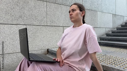 Young woman in pink shirt sitting on stairs with laptop, looking away. Urban setting with stone steps. Remote work, contemplation, lifestyle