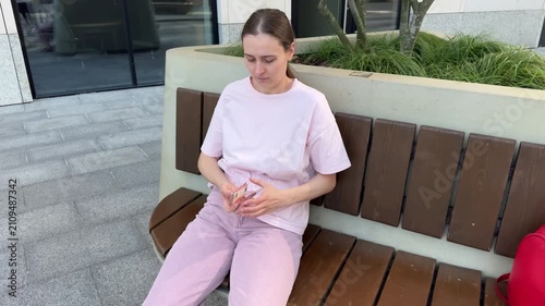 Young woman in pink clothes sitting on outdoor bench taking euro banknotes from pocket and counting them. Urban setting with modern building. Money handling, finance, payment