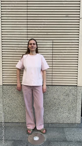 Young woman in pink casual outfit standing with hands on hips against modern slatted wall. Full body portrait, confident pose. Urban setting, contemporary fashion, lifestyle