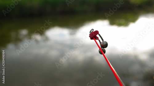Bell bite indicator attached to fishing rod moving after fish strike near waterside. Concept of sport fishing and freshwater angling