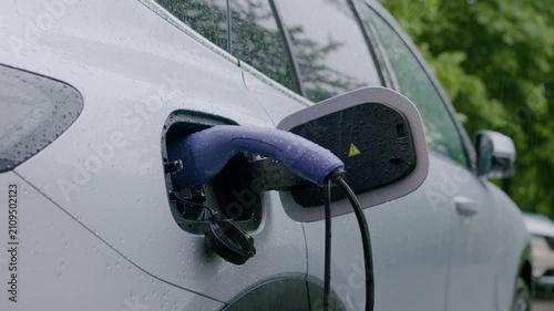 Electric vehicle charging in the rain with a close-up view of the charging port and cable, droplets of water visible on the car's surface and surrounding greenery