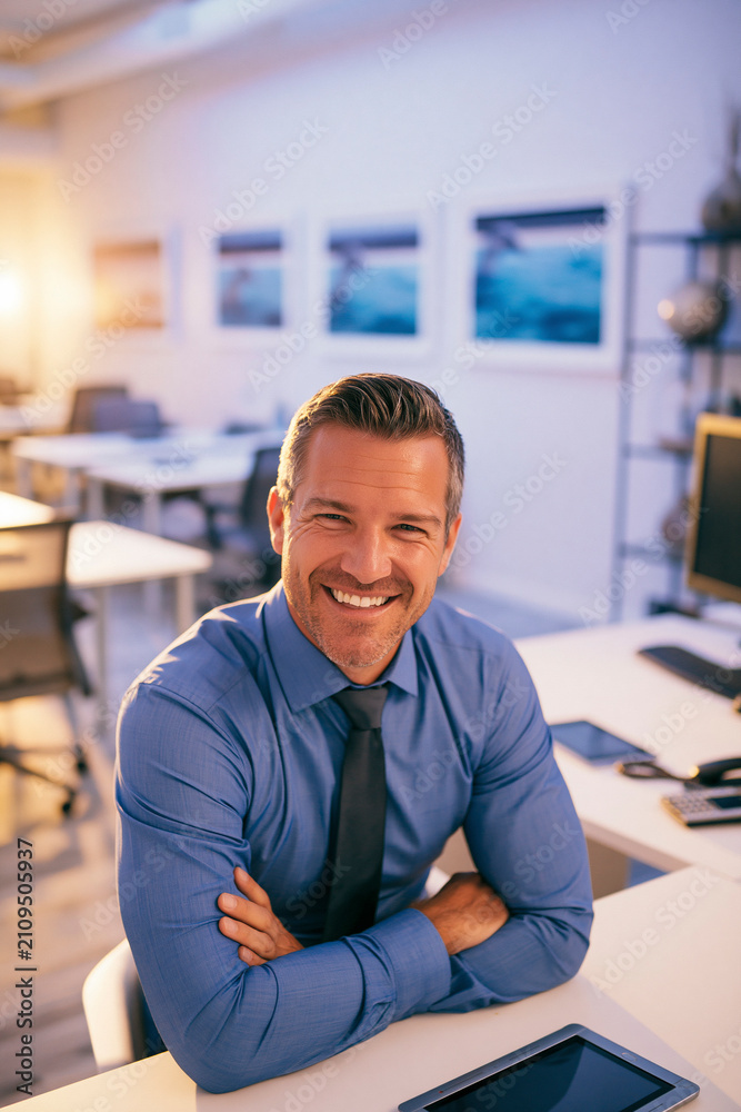 Obraz premium A man with dark hair and a blue shirt with a dark patterned tie, resting his hands behind his head and smiling, sitting at a white desk.