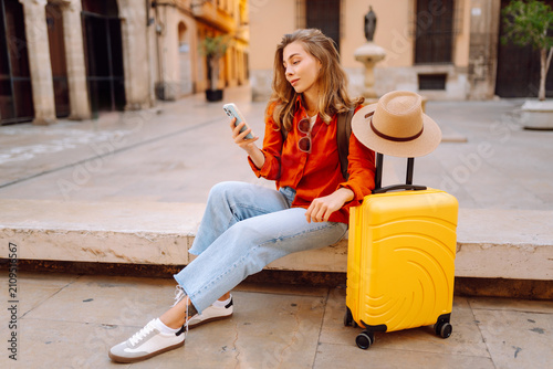 Tourist wearing hat and carrying a brightly colored suitcase sits in town square, holding phone. A young woman with a smartphone is relaxing outdoors. Concept for tourism, weekends, or vacations.