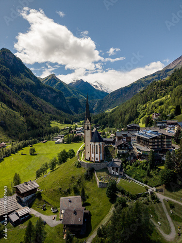 Hof, Austria - June 11 2026: Aerial drone footage of Pfarrkirche Heiligenblut, church in Hof, Austria. Beautiful weather with snowy mountains in the background. Heaven in the Austrian Alps. Grossglock