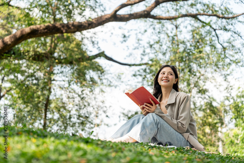 Young asian woman relaxing reading book sitting at public park life style outdoor. Happy female Take a break enjoy time living in the nature. Human and nature together