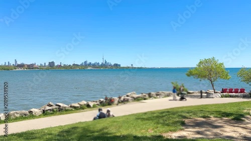 Toronto skyline across Lake Ontario from Sheldon Lookout. People enjoy a sunny summer day relaxing in the green waterfront park. Bicycles rest near the paved promenade under the clear blue sky.