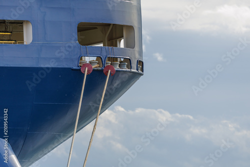 Close-up of a RORO ship's stern with heavy mooring lines running through a chock opening with roller fairleads and secured with red rat guards, set against a cloudy blue sky.