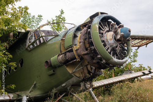 Weathered An-2 military plane abandoned in overgrown field. Aircraft propeller and fuselage with signs of decay under cloudy sky