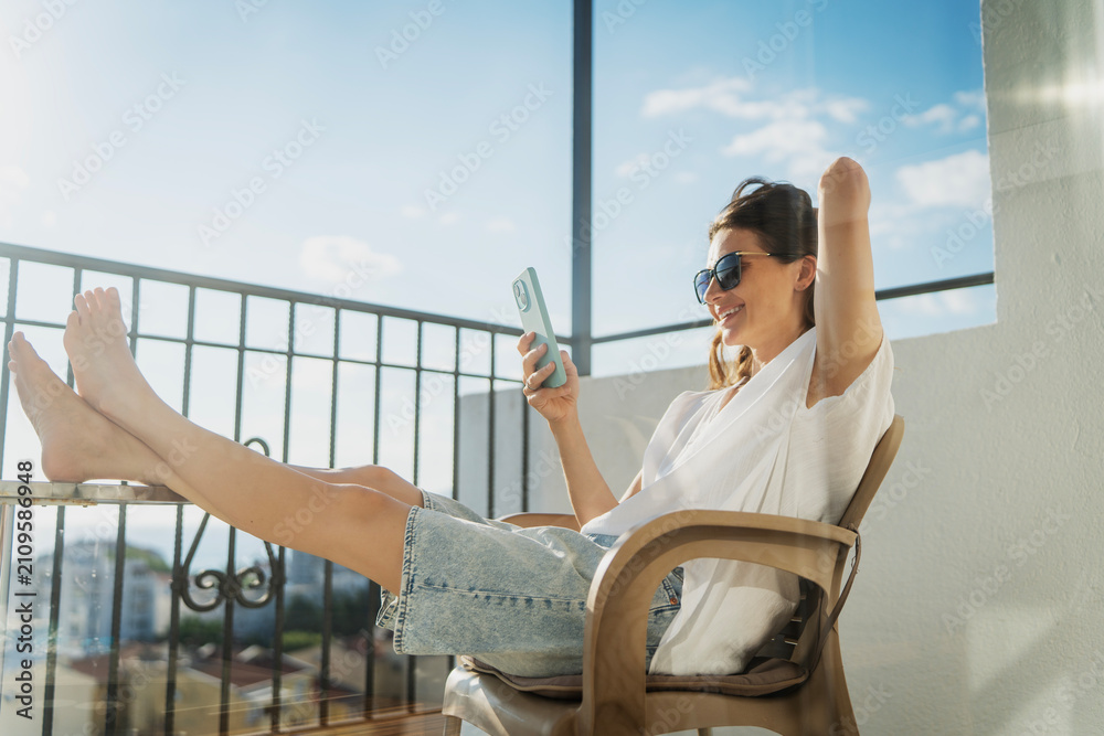 Obraz premium Happy young woman sitting on balcony chair with barefoot legs on table and looking at smartphone screen.