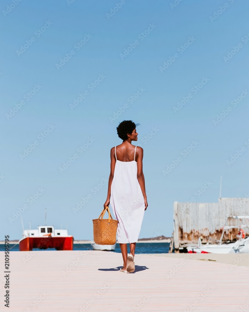 Fototapeta premium Black woman walking on a sun-drenched wooden dock with a straw bag for nautical style and summer travel concepts, plenty of copy space