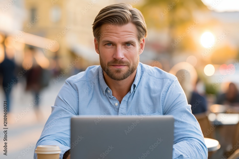 Fototapeta premium A handsome man with beard and blue shirt working on a laptop outdoors during golden hour with bokeh background.