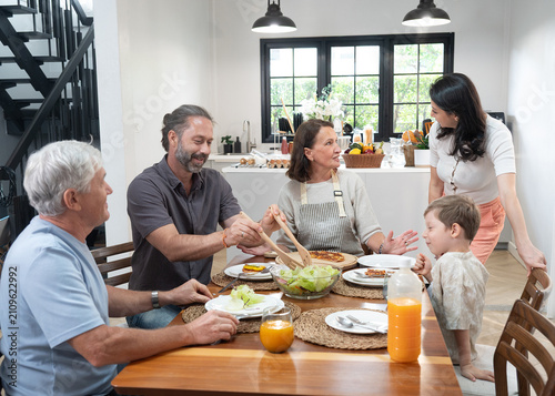 Multigenerational family gathers around kitchen table sharing meal conversation and warm smiles with salad juice and pancakes in bright modern dining area creating cozy home atmosphere