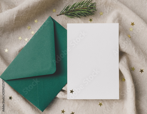 Empty white greeting card and open dark green envelope with golden star confetti and pine needles on a neutral textured textile, perfect for winter holiday mockups