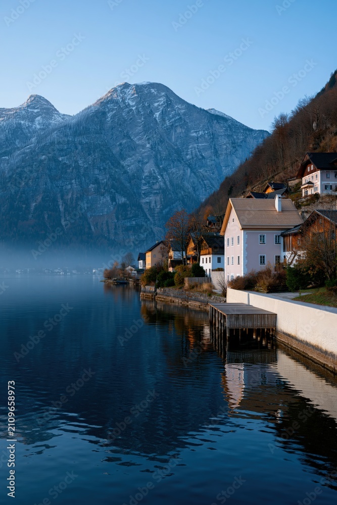 Fototapeta premium Serene waterfront houses by a reflective mountain lake with clear sky and soft morning light for background and travel banner design