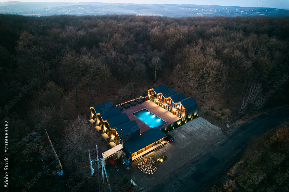 Obraz premium Aerial night view shows modern resort cabins around glowing pool. Warm lights, spacious wooden terrace and secluded forest setting create inviting luxury retreat concept