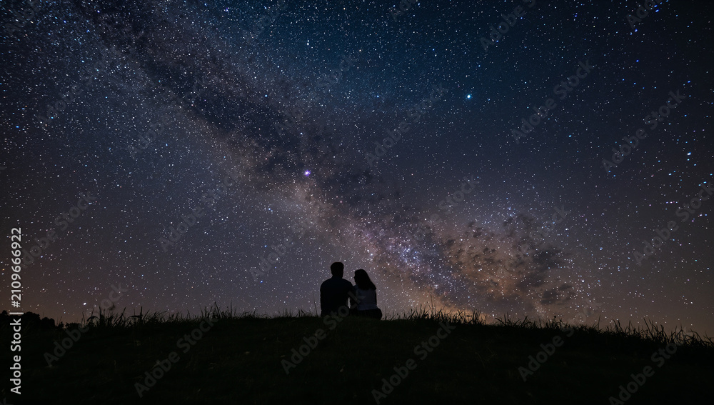 Fototapeta premium 壮大な天の川が広がる夜空の満天の星々を草の丘の上に並んで座り見上げる二人のシルエット