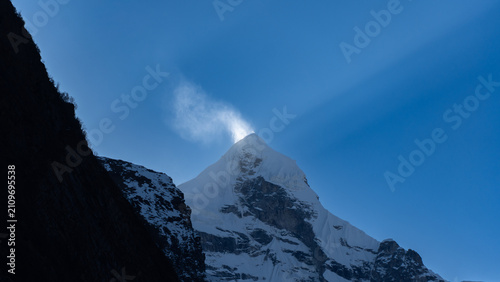 A majestic snow-capped mountain peak with a bird perched on its summit against a clear blue sky and rocky terrain in the foreground
