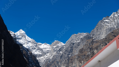 Snow-capped Neelkanth mountain peak stands tall against a clear blue sky, framed by rugged rock formations and partially visible under a bright sun with rays of light shining down.