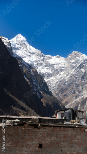 Neelkanth peak rises majestically against a clear blue sky, with rugged rock formations and patches of snow. The scene is framed by rick houses with metal roofs, a large water tank on its roof