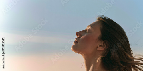 Young woman enjoying fresh air outdoors at sunset. Female model inhaling deeply with eyes closed in serene natural light.