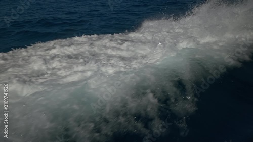 Turbulent white foam water trail behind a large boat in a calm blue sea on a sunny day, slow motion