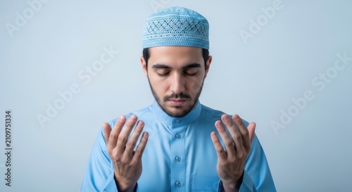 Young muslim man wearing blue traditional clothes praying with hands raised in humble devotion
