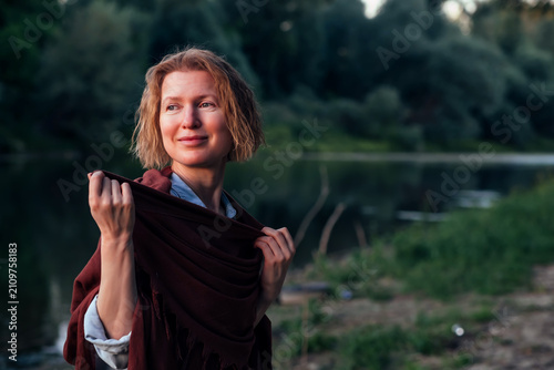 Quadro su tela Peaceful woman wrapped in a warm shawl by a quiet river at sunset, enjoying a serene moment in nature