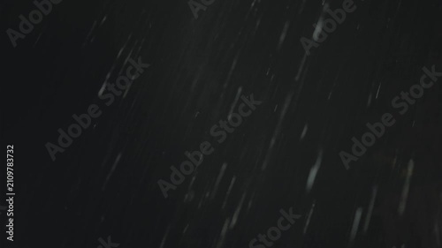 Heavy diagonal rain streaks glow against the blue grey night sky above a dark treeline, a passing flash briefly lights the downpour over the rural countryside