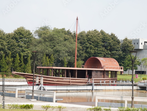 A traditional wooden ship replica with a tall red mast sits stationary in a serene park setting during the day.