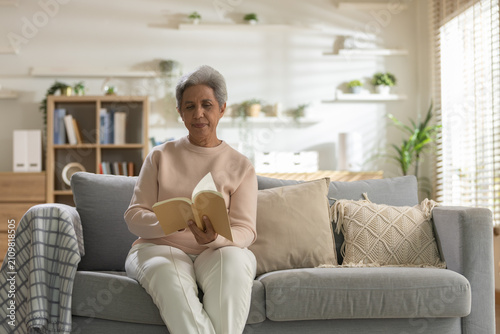 Fotografie Senior woman reading a book while relaxing on a cozy sofa at home, representing lifelong learning, peaceful retirement, healthy aging, mindfulness, home comfort, quiet leisure, wellness lifestyle
