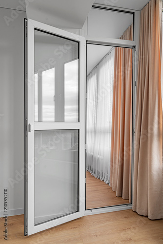 An open white plastic glass door leads to a modern balcony interior with curtains and wooden laminate floor.