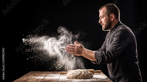 Super slow motion close up of professional artisan baker chef sprinkles flour on raw loaf of dough while making homemade bread, pasta or pizza on rustic wooden table in traditional bakery kitchen