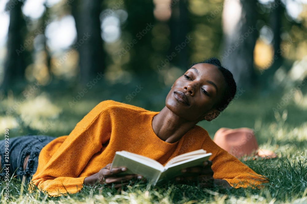 Fototapeta premium Woman reading book outdoors lying on grass in orange sweater for cozy lifestyle background and mindful relaxation banner