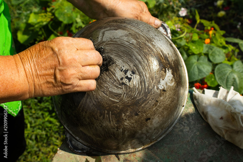 shot of an elderly woman's hands using a steel wool scrubber to clean soot and grease off the bottom of an inverted metal cauldron or kazan outdoors.