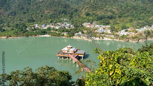 A serene view of Dhari Devi Temple on a riverbank with a suspension bridge stretching across the tranquil Ganges in Rudraprayag, surrounded by green hills and vibrant village houses.