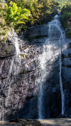 A majestic waterfall cascading down a rugged mountain cliff surrounded by lush greenery under bright sunlight with clear blue skies, creating a serene and adventurous atmosphere