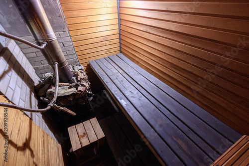 wooden sauna interior features horizontal plank walls and ceiling, a built‑in bench, a stone‑filled heater with a metal chimney, and warm ambient lighting