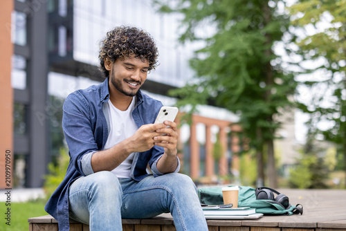 Young man with curly hair smiling while using a smartphone. Relaxing outdoors on campus with a backpack. Books. Headphones. And coffee cup beside him